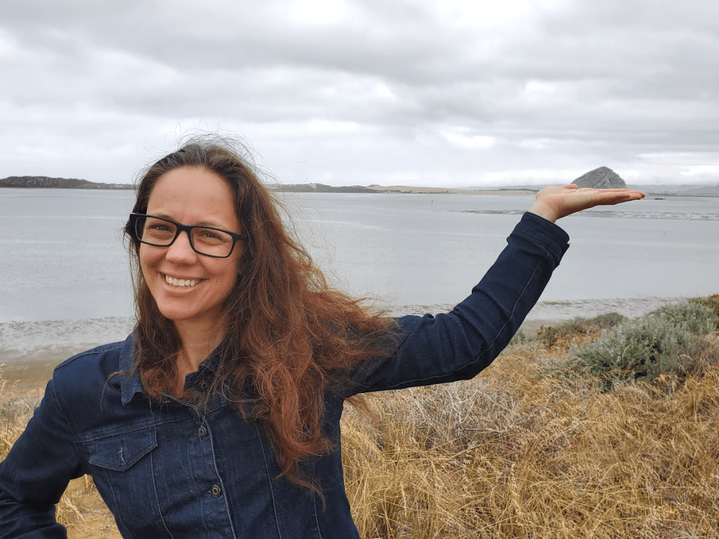 Shannon standing next to a lake in Los Osos California in 2019. She has her hand upturned on the horizon so it looks like she's holding up a distant hill