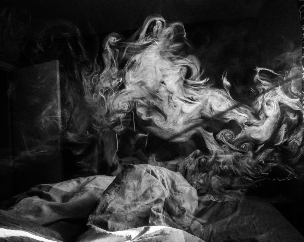 Black and white photo of a dark room,  silhouetted figure in a dress sitting on a bed, white smoke swirling all around