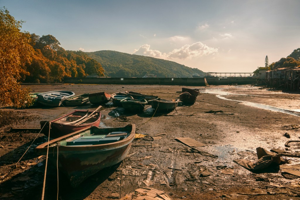 Color photo of several rowboats grounded on a dry, brown riverbed with a tiny bit of muddy water nearby.