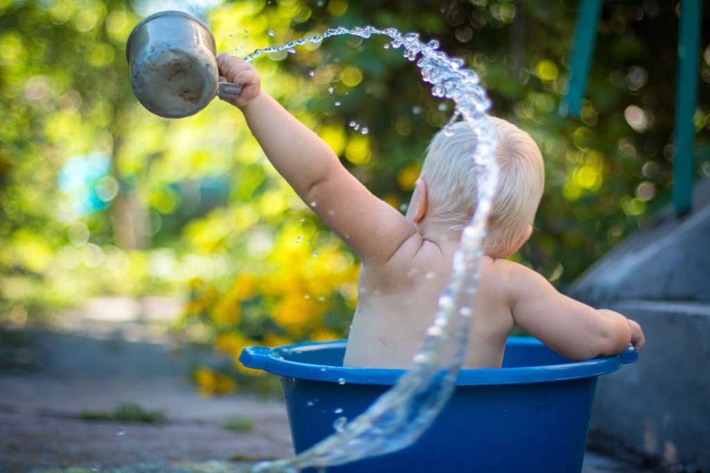 Color photo of the back of a baby in a blue washtub flinging a metal cup of water in an arc.