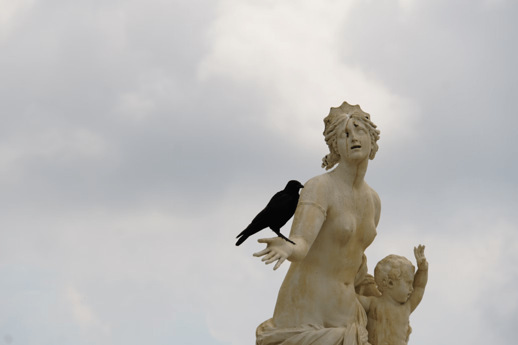 Photo by Jan Zinnbauer on Unsplash. Marble statue of a woman and child outside under a cloudy sky (Latona's fountain in Versailles). Woman's arm is outstretched and a live crow is perched on her wrist. 