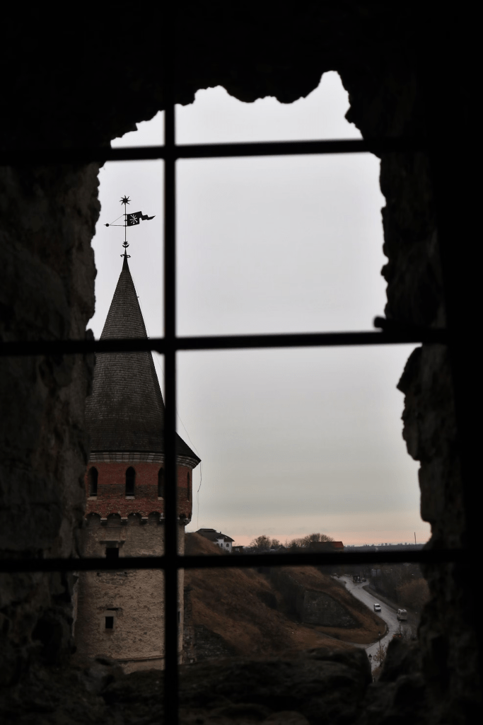 Photo by Yevheniia on Unsplash. Picture from inside a barred tower window with jagged edges looking out over a dark, adjacent tower and the countryside below at dusk. 