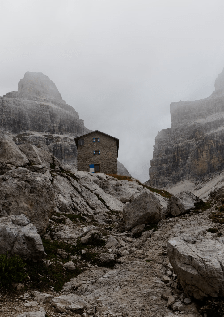 Photo by Wilfried Santer on Unsplash. Image of a rocky, hilly stretch of mountains against a foggy sky with a single stone hut perched precariously on the rocks. 