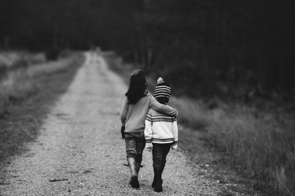 Photo by Annie Spratt on Unsplash. Black and white image of two young children walking away from the camera down an unpaved road. The taller one has an arm around the shorter one's shoulders. 